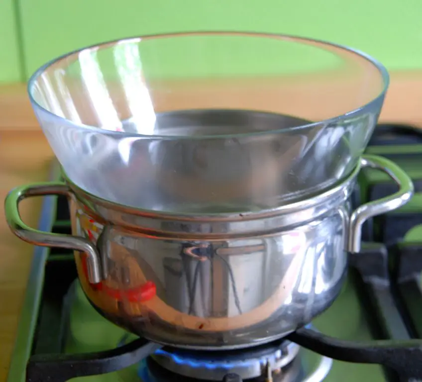 a person assembling a homemade double boiler by placing a metal bowl over a saucepan filled with water.