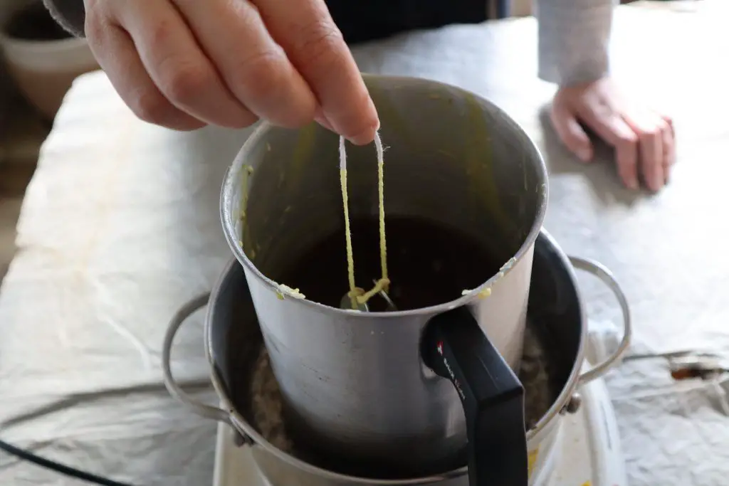 a person dipping a candle wick into melted beeswax