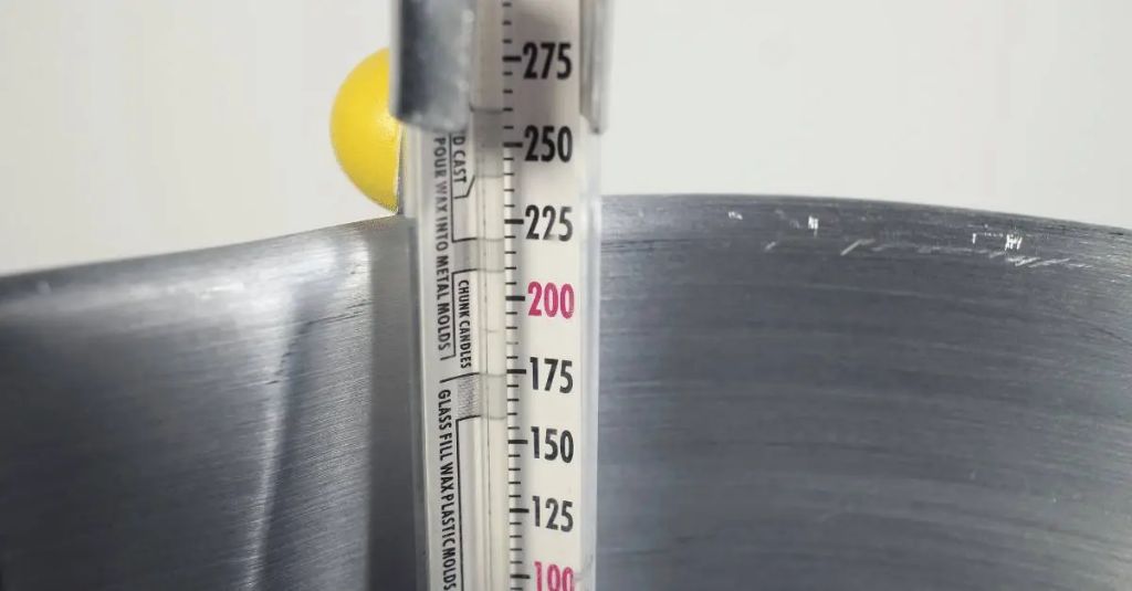 a person holding a thermometer in a melting pot of candle wax.