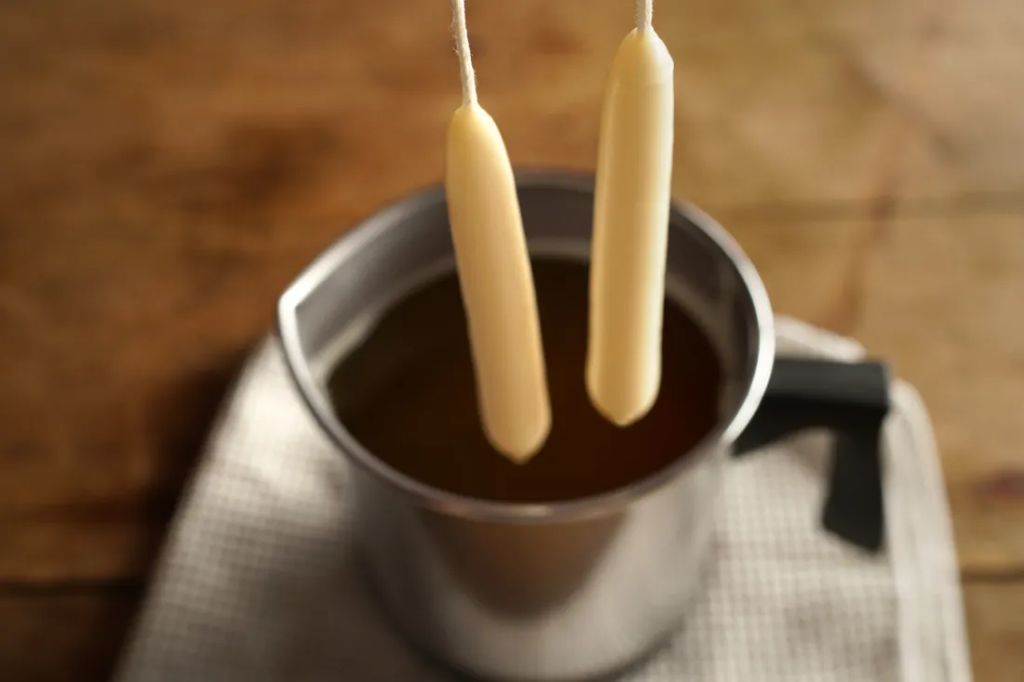 a person holding a wick and slowly dipping it into a pot of melted wax to create a candle.