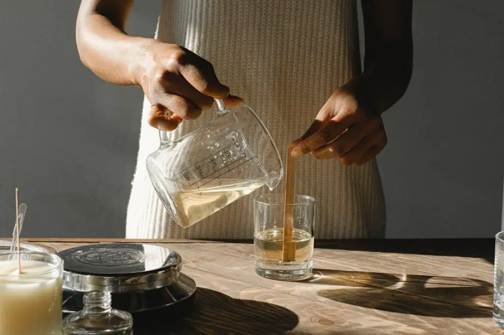 a person wearing a protective mask and gloves while pouring melted wax into a candle mold.